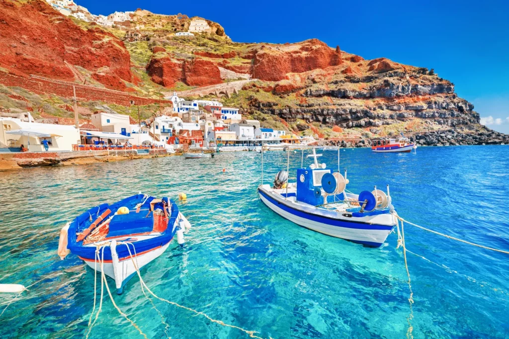 Hellas. Et fantastisk vakkert landskap med to fiskebåter som ligger ved kai i fascinerende blått vann ved det fantastiske gamle havnepanoramaet i landsbyen Oia Ia på den greske øya Santorini i Egeerhavet.