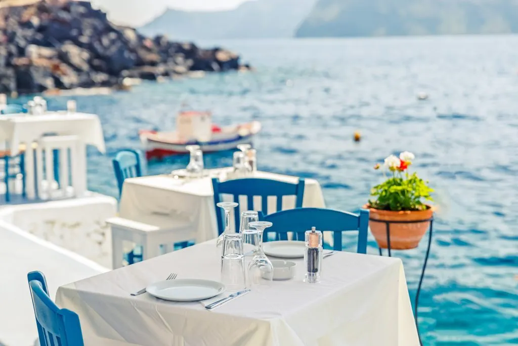 Griechenland, Santorini. Restaurant mit serviert Tisch in Meeresfront der Ägäis auf Santorini Kykladen Insel mit atemberaubenden, erstaunlichen und unglaublichen Blick auf das Wasser und die Böschung von Oia Ia Dorf.